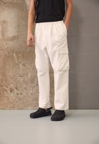 White cargo pants with side pockets and an elastic waistband, paired with black hole-patterned clogs on a light gray surface.