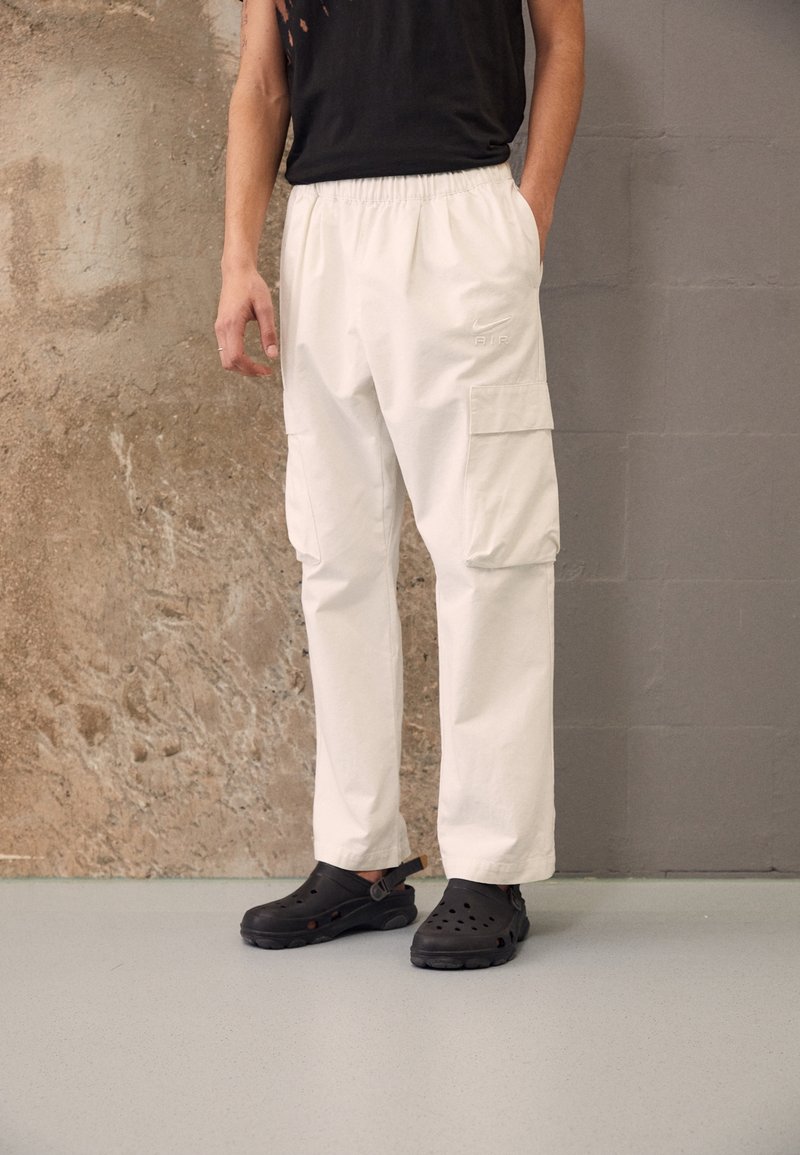 White cargo pants with side pockets and an elastic waistband, paired with black hole-patterned clogs on a light gray surface.