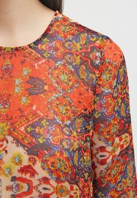 Blusa a maniche corte rossa e arancione con una texture trasparente. Presenta disegni intricati floreali e geometrici con accenti blu e gialli. Collo rotondo.