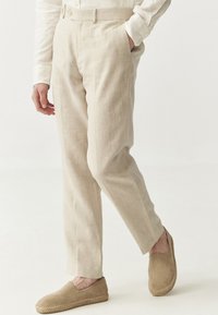 Beige Leinenhosen mit geradem Schnitt, Fronttaschen und Knopfverschluss, kombiniert mit hellbraunen Espadrilles.