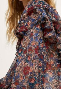 Femme portant un chemisier transparent avec des détails froncés aux épaules et un motif floral et paisley multicolore dans des tons de rouge, bleu et beige.