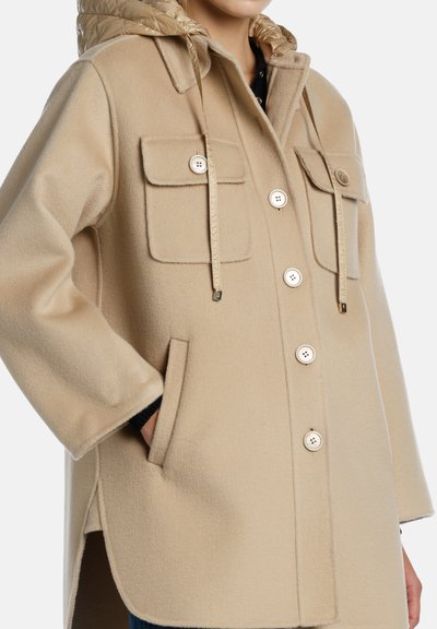 Cappotto in lana color beige con cappuccio trapuntato, grandi tasche anteriori, chiusura con bottoni dorati e spacco laterale per facilitare il movimento.