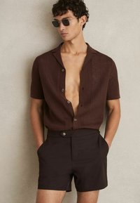 Cămașă scurtă, cu mâneci scurte, din tricot deschis, de culoare maronie, cu butoane, purtată cu pantaloni scurți maro închis care au un buton lateral. Textură netedă, croială casual.
