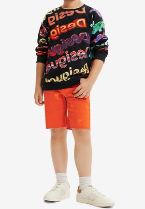 Sweatshirt noir avec texte multicolore, associé à un short en denim orange avec un ourlet effiloché. Baskets blanches avec des accents clairs et beige.