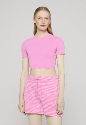 Nainen seisoo vaaleaa taustaa vasten, pukeutuneena vaaleanpunaiseen, lyhytmidrattu T-paitaan ja vaaleanpunaisiin kiristysnyörillisiin shortsseihin, joissa on seepra-kuvio.