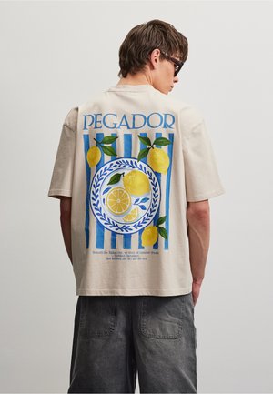 Mand iført beige t-shirt med blå striber, citroner og teksten "PEGADOR" på ryggen, kombineret med mørkegrå bukser og sorte solbriller.
