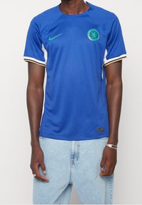 Camiseta de fútbol Nike azul de mangas cortas, con cuello redondo, logo de Chelsea en verde y rayas de acento en blanco y dorado.