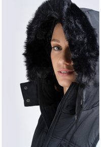 Schwarze Kapuzenjacke mit Kunstfellbesatz, gestepptem Design und Reißverschluss. Verfügt über metallene Druckknöpfe im Kragenbereich.