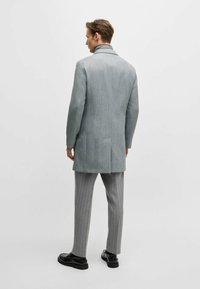 Ljusgrå skräddarsydd kappa med en strukturerad krage, en enda vent, och sidofickor, i kombination med grå ränderbyxor och svarta skor.