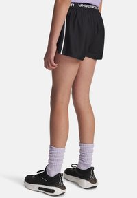 En person i svarta träningsshorts med vit rand, lavendelfärgade strumpor och svarta sneakers, står mot en vit bakgrund.