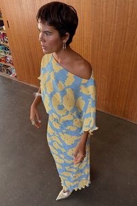 Modré a žluté šaty s vzorem, plisováním a odhalenými rameny. Mají volné rukávy a sahají až ke kotníkům s texturovaným vzhledem.