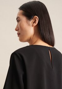 Top nero con scollatura rotonda, maniche corte e apertura a chiave sul retro fissata con un bottone. Tessuto liscio con finitura opaca.