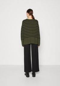 Übergroßer olivgrüner Pullover mit schwarzen Streifen, langen Ärmeln, geripptem Saum, kombiniert mit weiten schwarzen Hosen und klobigen schwarzen Boots.