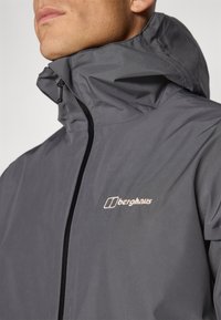 Gray waterproof jacket with high collar, zip closure, and Berghaus logo in light-colored embroidery. Smooth texture, minimalistic design.