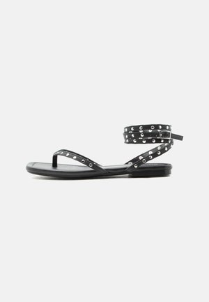 Sort flat sandal med tynne stropper, sølv nagler og en ankelrem med spenne på en ensfarget lys bakgrunn.
