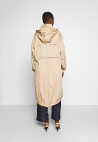 Henkilö, joka on pukeutunut beigeen huppariin, pitkä takki, tummiin lyhyisiin housuihin ja valkoisiin korkokenkiin, seisoo yksinkertaista vaaleanharmaata taustaa vasten.