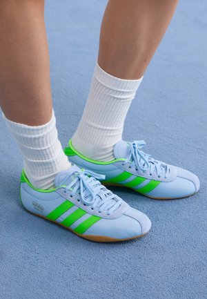 Világoskék sportcipők zöld részletekkel és három csíkkal. Simább textúrával és gumitalppal rendelkeznek. Fehér bordázott zoknikkal viselve.