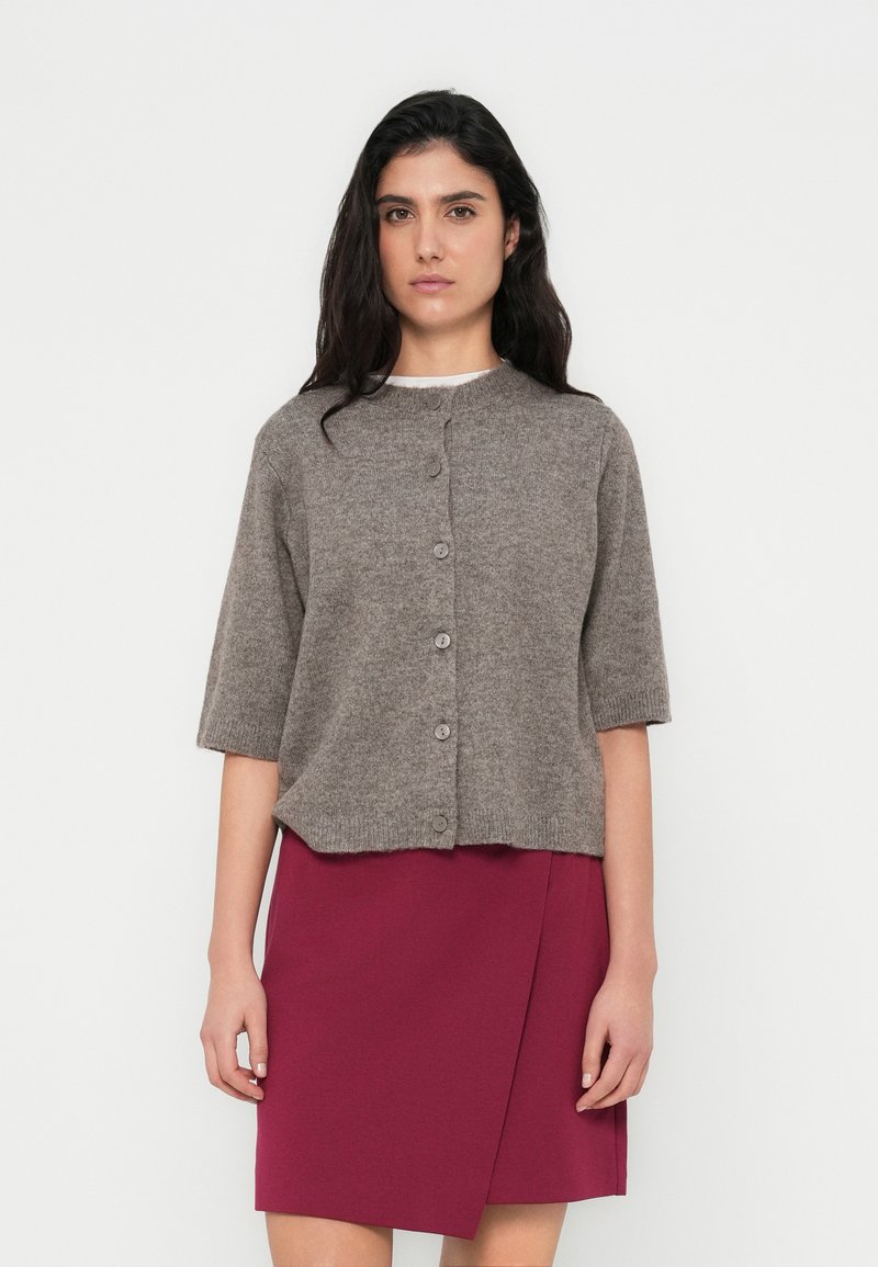 Γκρι πλεκτή ζακέτα με κοντά μανίκια, στρογγυλή λαιμόκοψη και έξι κουμπιά. Συνδυάζεται με μπορντό φούστα τύπου wrap. Λεία υφή, καθημερινό σχέδιο.