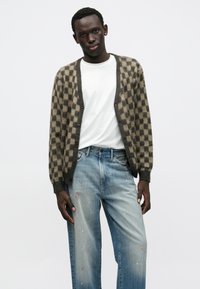 Καρντιγκάν σε καφέ και μπεζ καρό μοτίβο, κατασκευασμένο από μαλακό υλικό, με κουμπιά μπροστά και ριμπ μανσέτες. Φορεμένο πάνω από ένα λευκό πουκάμισο και συνδυασμένο με ανοιχτό μπλε τζιν με σκισίματα.