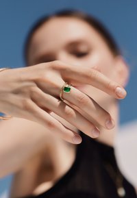 Anillos de oro con una piedra preciosa verde en forma de cuadrado, apilados con una banda texturizada, sostenidos por una mano contra un cielo azul claro.