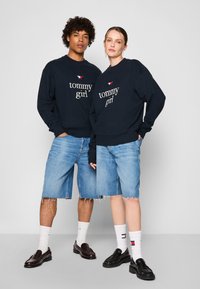 Marineblå sweatshirts med "tommy girl" logo, parret med lyse denimshorts med frynset nederkant og sorte loafers. Hvide sokker med logoer.