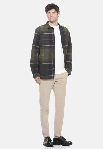 Camicia in flanella a quadri nei toni del verde scuro, grigio e burgundy. Chiusura a bottoni, vestibilità comoda. Abbinata a pantaloni beige e scarpe nere.