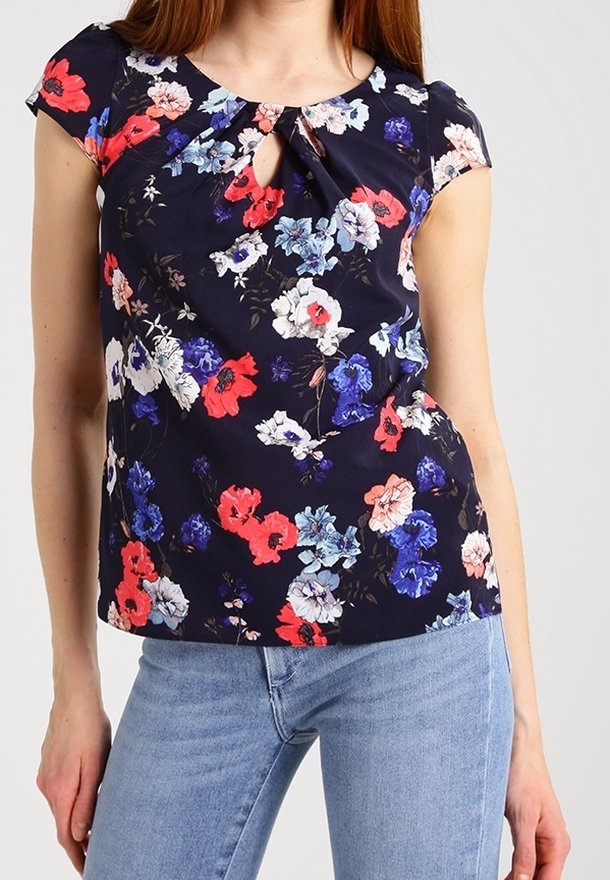Blus med blommönster på en marin bakgrund, som har capärmar, en nyckelhålsknappning vid halsen och livfulla röda, blå och vita blommor.