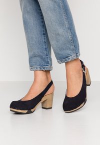 Svarta mocka slingback högklackade skor med rund tå, träblockklack och kontrasterande beige sula, prydda med dekorativa nitar längs kanten.