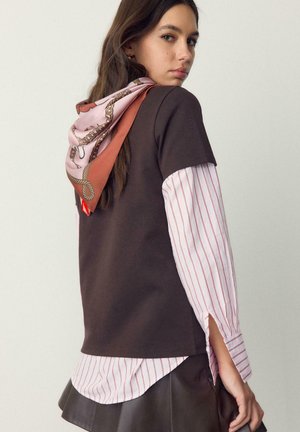 Giovane donna con indosso una maglia marrone a strati sopra una camicia a righe, gonna di pelle marrone e sciarpa rosa fantasia annodata dietro, che guarda di lato.