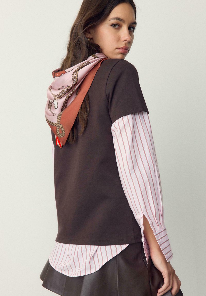 Jeune femme portant un haut marron superposé sur une chemise rayée, une jupe en cuir marron et une écharpe rose à motifs nouée à l'arrière, regardant de côté.
