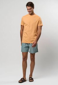 Gaisdziemā apelsīnu krāsas kokvilnas T-kreklā ar mazu zilu logotipu, kombinācijā ar zilām un baltām svītrām veidotiem šortiem. Valkā ar tumši brūnām čībiņām.