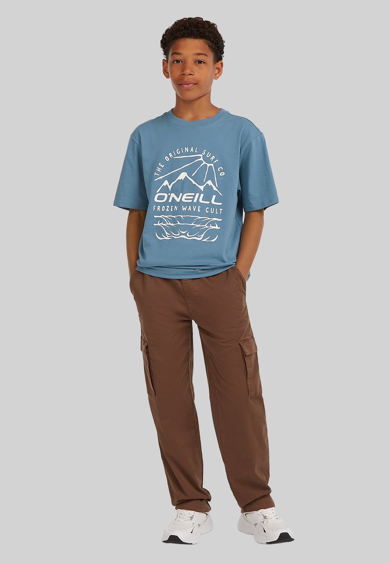 Camiseta de algodón azul con diseño gráfico blanco de montañas y olas. Pantalones cargo marrones con bolsillos laterales, combinados con zapatillas blancas.