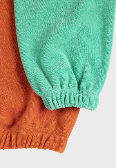 Deux poignets de manche en jersey éponge élastiqué, l'un turquoise et l'autre orange rouille, se chevauchant sur un fond clair.