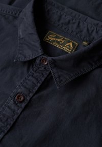 Camisa de botones azul marino hecha de algodón con una textura suave. Presenta un cuello clásico, botones marrones y una etiqueta que dice "Superdry Utility Wear."
