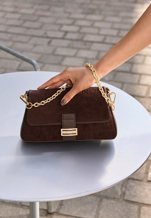 Ръка с изрисувани нокти, държаща кафява чанта Valentino с златен верижен презрамка на кръгла бяла маса на открито.