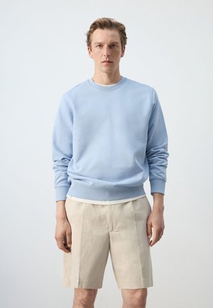 Muškarac u svijetloplom džemperu i bež kratkim hlačama do koljena stoji ispred jednolične svijetlosive pozadine.