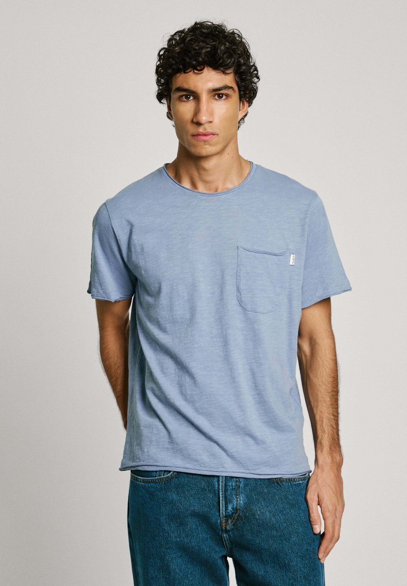 Šviesiai mėlynas medvilninis marškinėlis su apvaliu iškirpimu, trumpomis rankovėmis ir priekyje esančia kišenėle. Šiek tiek tekstūruota medžiaga su laisvu kirpimu.