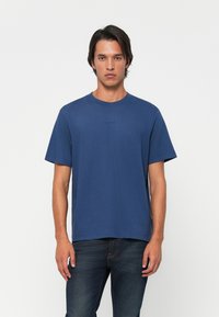 Young man standing with neutral expression wearing a plain blue short-sleeve t-shirt and dark jeans against a white background.