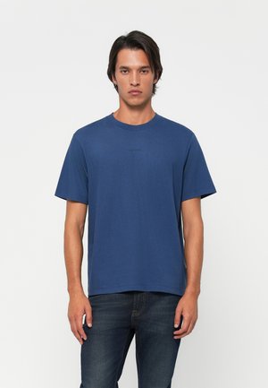 Junger Mann mit neutralem Ausdruck, der ein schlichtes blaues T-Shirt mit kurzen Ärmeln und dunkle Jeans vor einem weißen Hintergrund trägt.