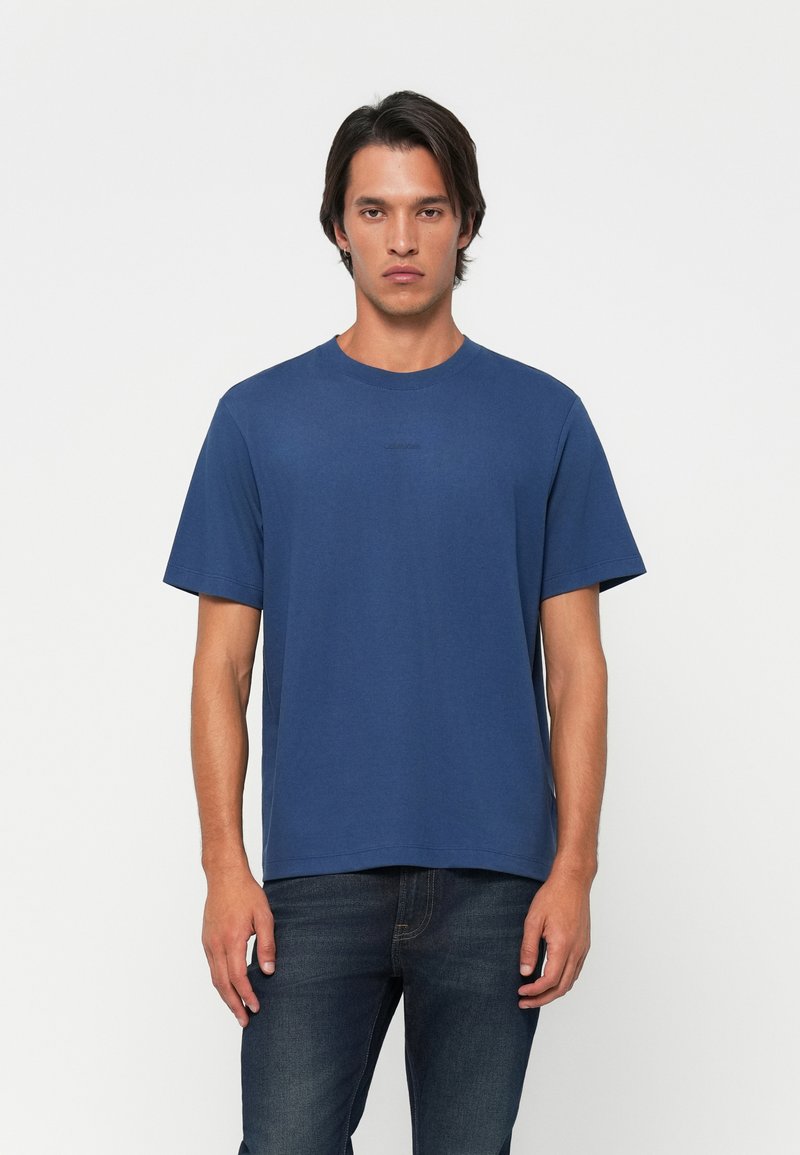 Young man standing with neutral expression wearing a plain blue short-sleeve t-shirt and dark jeans against a white background.