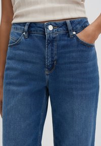 Donkerblauwe denim jeans hebben een hoge taille, voorzakken en zilveren hardware. De stof heeft een zachte textuur met minimale slijtage.