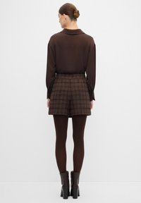Blusa marrone con colletto, abbinata a pantaloni corti a quadri marroni. La modella indossa collant scuri e stivali con tacco. Tessuto testurizzato con un motivo.