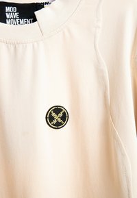 Camisa beige con parche circular negro y dorado que presenta las iniciales estilizadas "MWM" y el texto "MOD WAVE MOVEMENT" en el interior del cuello.