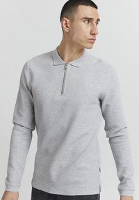 Graues, strukturiertes Langarm-Polo mit einem halben Reißverschluss am Kragen und seitlichen Schlitzen, das ein dezentes, gewebtes Muster und eine schlanke Passform aufweist.
