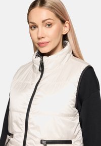 Lekka kremowa kamizelka puchowa z wysokim kołnierzem, detalami na suwak i czarnymi akcentami. Posiada wzorzystą podszewkę wewnętrzną oraz kieszenie na suwak.