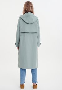 Long, light green raincoat with a hood, featuring a layered back design, cinched waist, and elastic cuffs, paired with blue jeans and brown shoes.