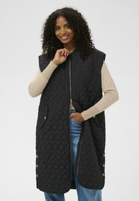 Veste matelassée noire avec fermeture éclair à l'avant, emmanchures surdimensionnées, poches latérales et motif en losange. Portée par-dessus un haut beige à manches longues.
