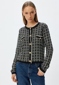 Cardigan noir et blanc à motifs avec un col ondulé, des boutons dorés et deux poches poitrine. Porté avec un jean en denim bleu.