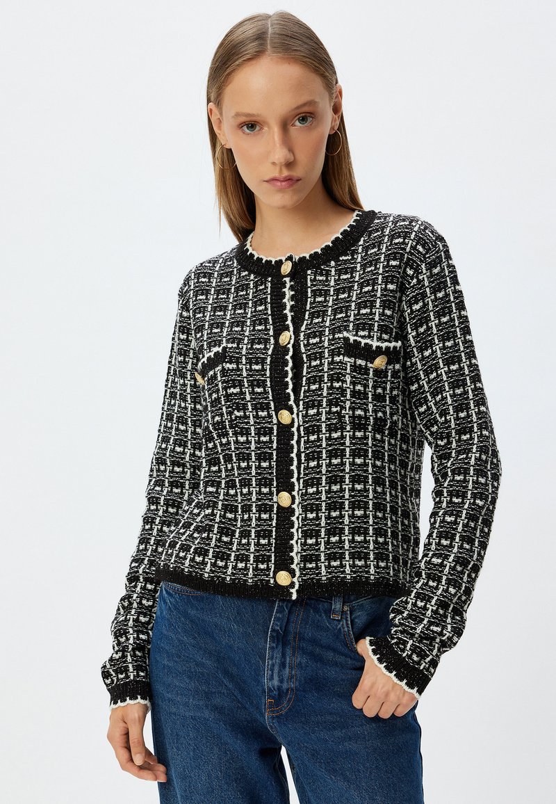 Cardigan noir et blanc à motifs avec un col ondulé, des boutons dorés et deux poches poitrine. Porté avec un jean en denim bleu.