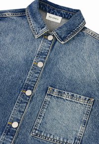 Denimskjorte med spiss krage, sølvfargede knappelukkinger, frontlomme og kontrasterende gul tråd på en falmet blå stoff.